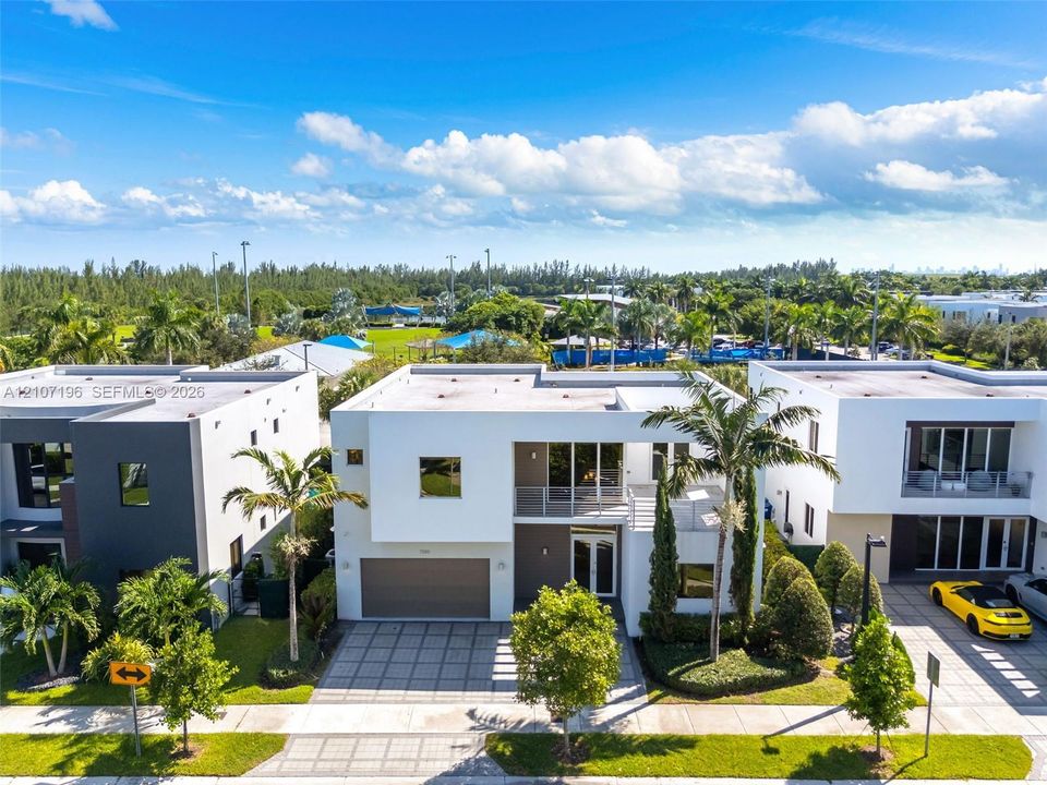 Modern two-story architectural exterior featuring a flat roofline, grid-patterned concrete paver driveway, attached two-car garage, second-level balcony with metal railing, and expansive glass window wall