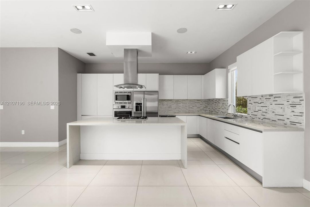 Kitchen featuring flat-panel cabinetry, a central island with a waterfall countertop, stainless steel appliances, a linear-patterned backsplash, and light-toned tile flooring