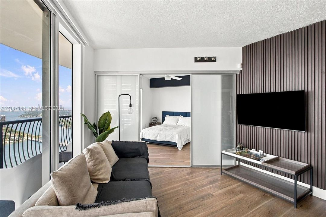 Floor-to-ceiling windows, wood-finish flooring, accent wall with vertical slat detailing, sliding glass room divider, and private balcony with water exposure