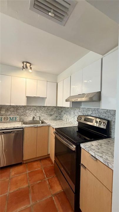 Kitchen featuring high-gloss white upper cabinetry, wood-finish base cabinetry, stainless steel appliances, stone-patterned countertops, and terracotta tile flooring