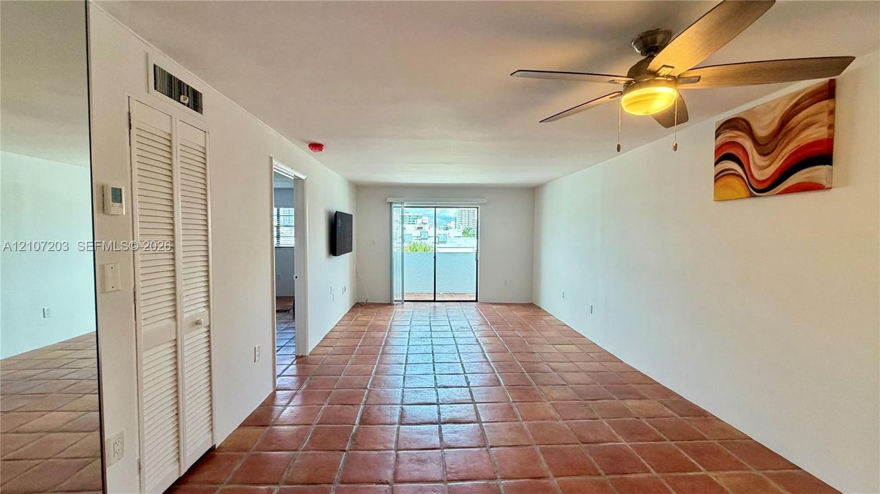 Large open-concept room featuring terra cotta tile flooring, floor-to-ceiling mirror paneling, a ceiling fan with integrated lighting, and sliding glass doors