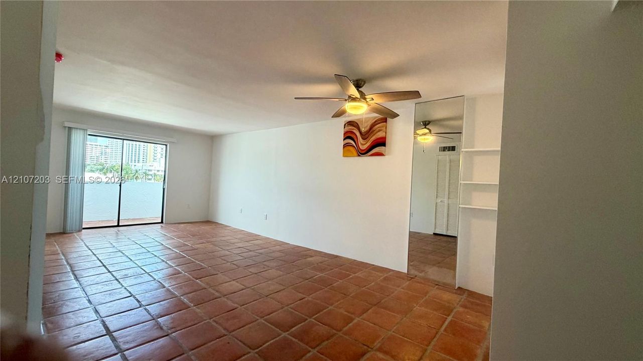 Open-concept interior featuring terracotta tile flooring, a ceiling fan with integrated lighting, full-height wall-mounted mirror, and sliding glass doors with vertical blinds