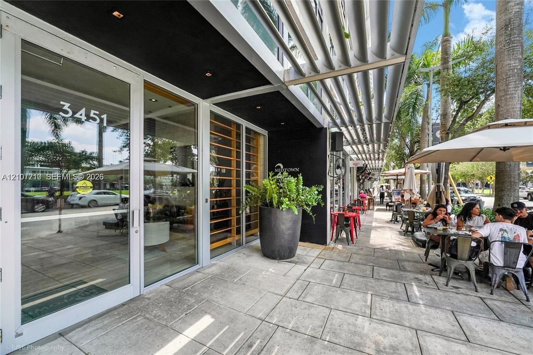 Exterior entrance featuring floor-to-ceiling glass paneling, overhead metal louvered canopy, paved stone walkway, and recessed ceiling lighting