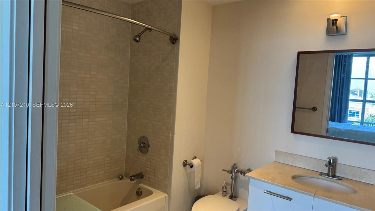 Bathroom featuring a shower-tub combination with light-toned mosaic wall tile, a vanity with a solid surface countertop and undermount sink, a wall-mounted mirror with a wood-finish frame, and polished metal hardware