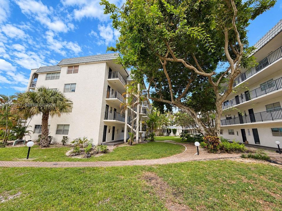 Multi-story buildings featuring exterior corridors with metal railings, brick-paved walkways, and established landscape including palm trees and mature shade trees
