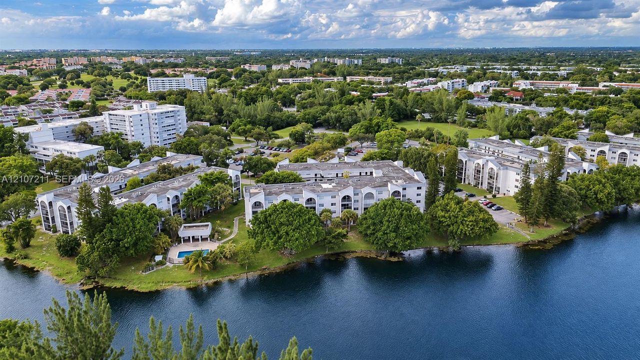 Aerial perspective of multi-story residential buildings featuring white stucco facades, arched balcony detailing, a waterfront location, mature trees, and an on-site swimming pool