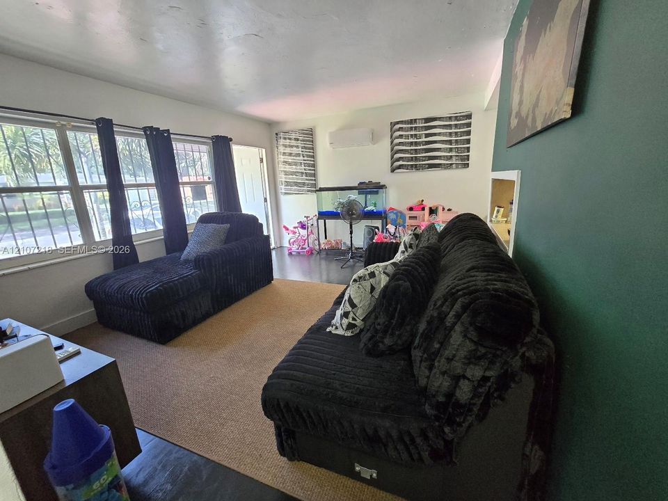 Living space featuring dark-toned flooring, wall-mounted climate control unit, large windows with horizontal blinds, and a built-in wall niche