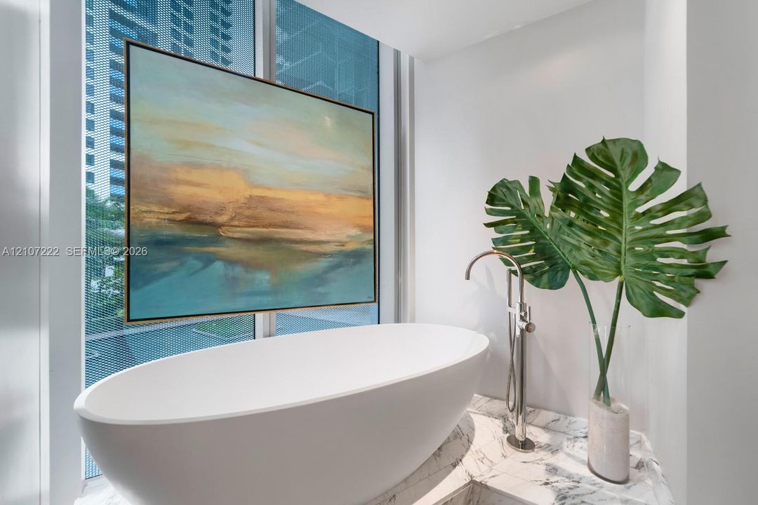 Freestanding white soaking tub with a floor-mounted metallic faucet, situated atop a stone-surfaced platform and flanked by floor-to-ceiling windows