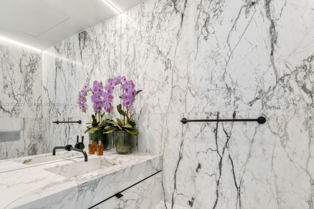 Full-height stone wall cladding featuring integrated cove lighting, vanity with matching stone countertop, undermount sink, and matte black wall-mounted fixtures