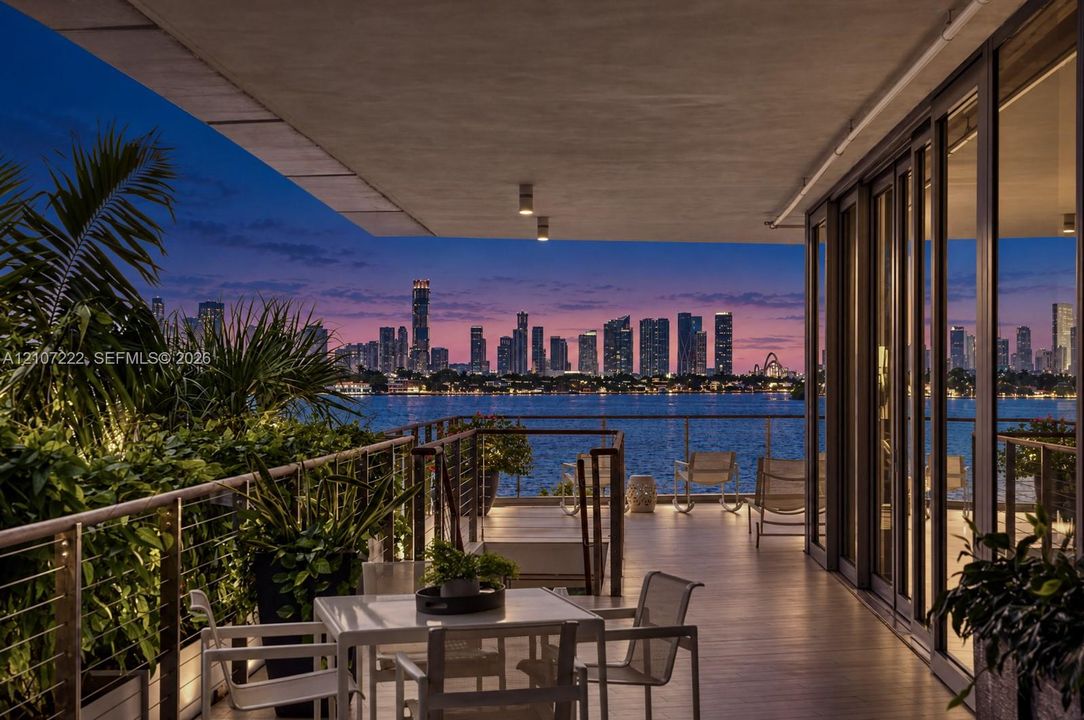 Expansive covered terrace featuring floor-to-ceiling glass walls, integrated ceiling lighting, and a metal cable railing system overlooking the water and city skyline