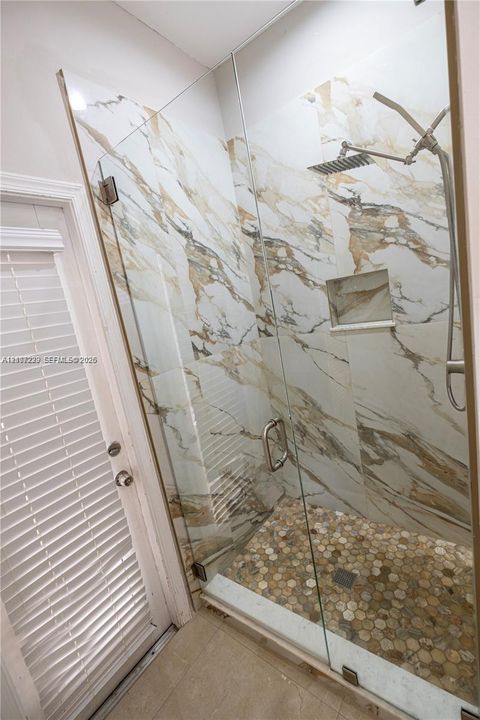 Walk-in shower featuring large-format stone-patterned tile walls, frameless glass enclosure, hexagonal mosaic tile flooring, built-in recessed wall niche, and rainfall shower head with handheld attachment