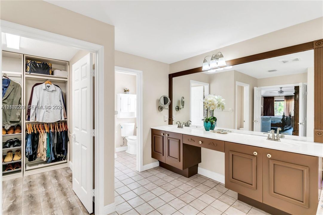 Dual-sink vanity with built-in cabinetry and stone-surface countertop, full-width mirror, tile flooring, and walk-in closet with integrated shelving