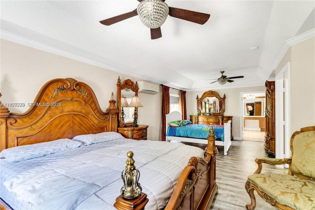 Tray ceiling featuring dual crystal-accented ceiling fans, wood-finish flooring, and a wall-mounted ductless climate control unit