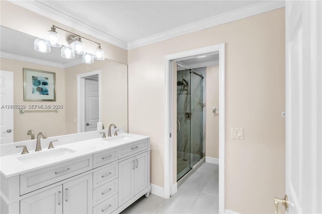 Double-sink vanity featuring stone-surfaced countertops, brushed nickel fixtures, crown molding, and a full-width mirror, accompanied by a glass-enclosed shower stall with tile flooring