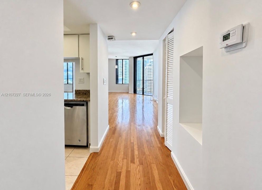 Hallway featuring wood-finish flooring, recessed lighting, and a built-in wall niche, with a partial view of a kitchen equipped with stainless steel dishwasher and stone-finish countertops