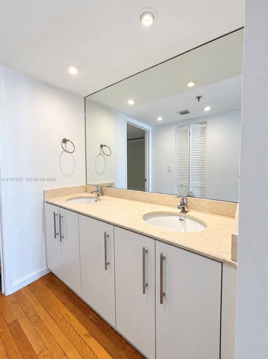 Dual-sink vanity featuring stone countertop, recessed oval basins, white cabinetry with brushed-metal pulls, and full-width wall mirror
