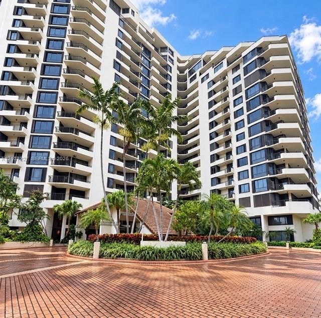 High-rise residential exterior featuring a multi-story facade, tiered private balconies, brick-paved circular motor court, and landscaped gardens with mature palm trees