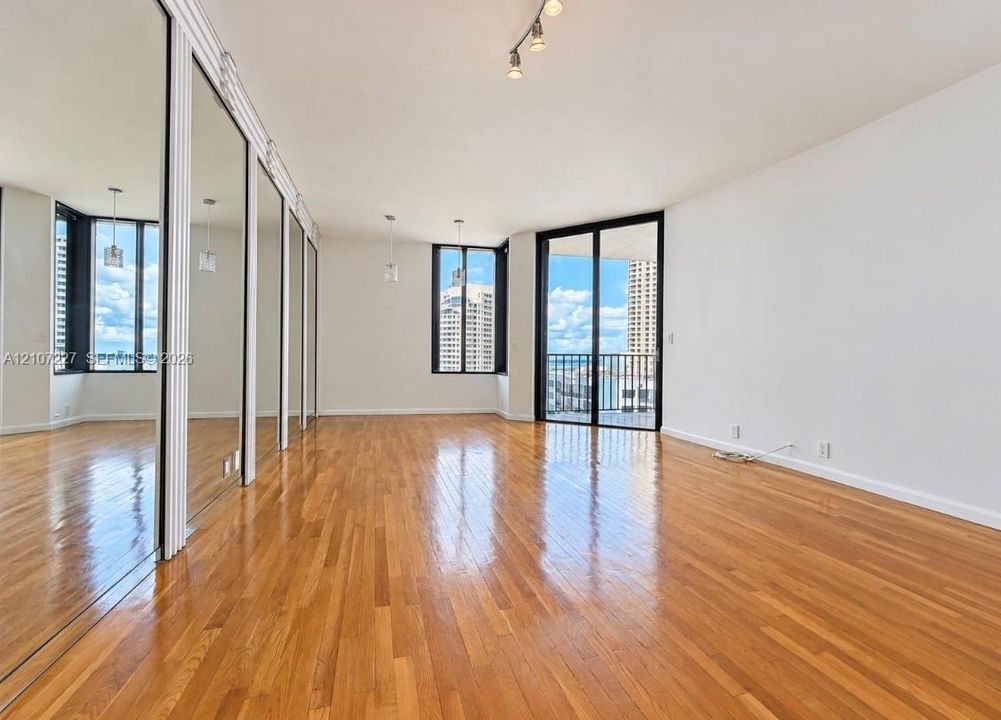 Wood-finish flooring, full-wall mirrored paneling, sliding glass door with balcony access, and multi-light track lighting fixtures