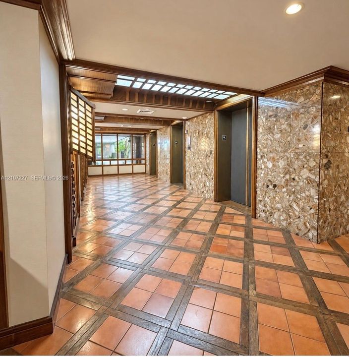 Common area featuring terracotta tile flooring with integrated wood trim, stone-clad feature walls, grid-patterned wood ceiling accents, and built-in elevator access