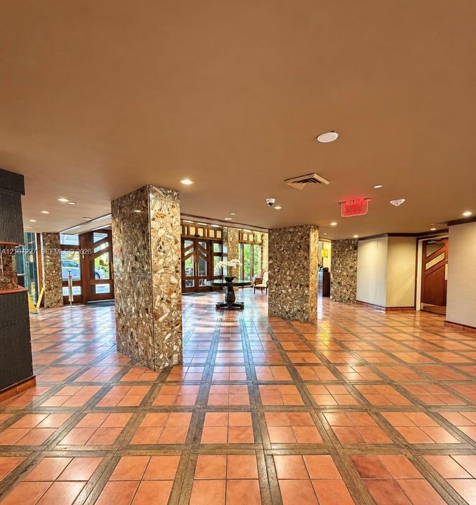 Lobby featuring terracotta tile flooring with inlay details, natural stone-clad structural pillars, recessed ceiling lighting, and multi-pane wood entry doors