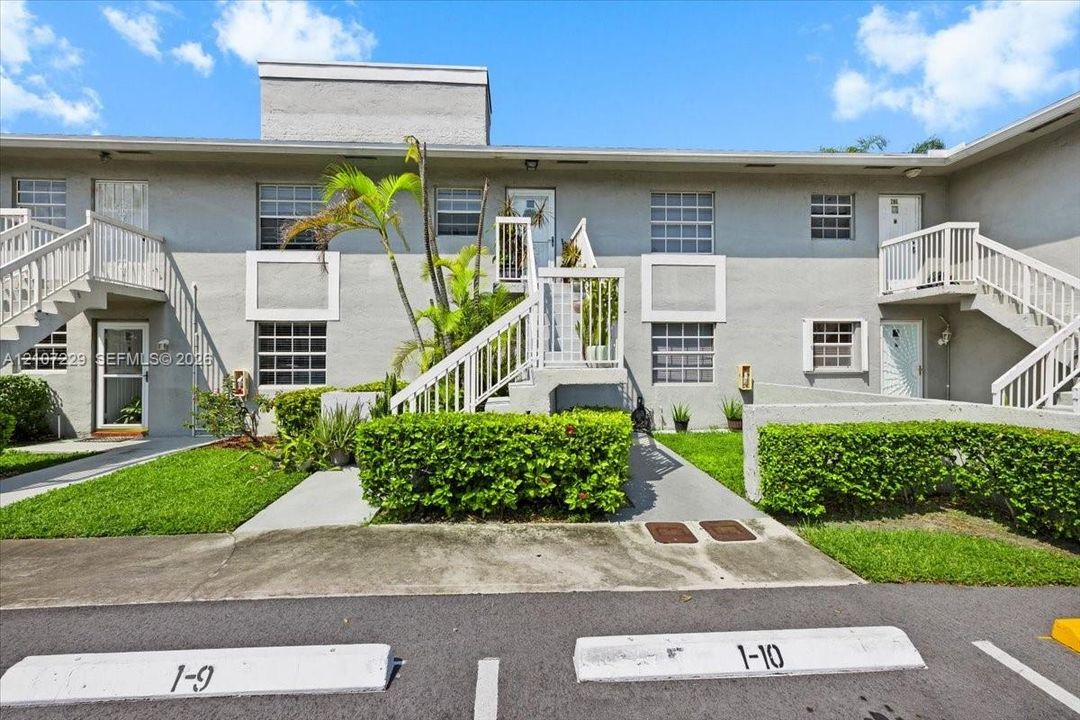 Two-story building exterior featuring smooth stucco siding, designated parking spaces, multi-level stairs with white railing, and manicured hedges