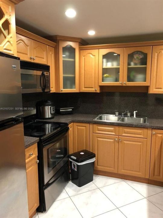 Kitchen featuring wood cabinetry with glass-panel upper doors, stone-surface countertops, stainless steel appliances, dual-basin sink with pull-down faucet, and tiled flooring