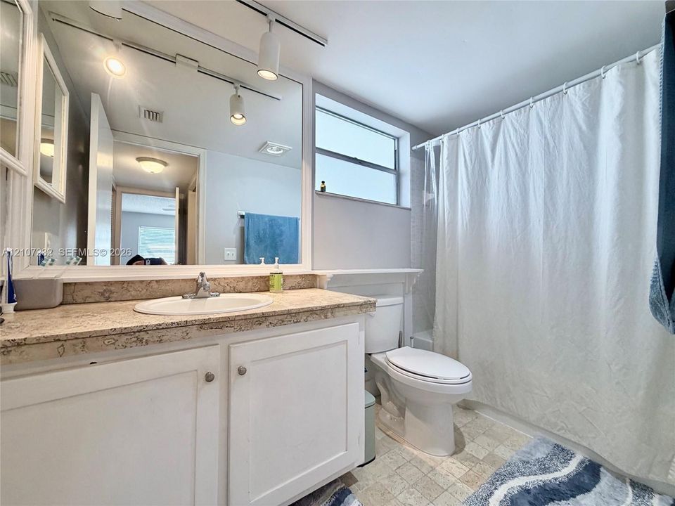 Bathroom featuring a stone-look countertop, integrated vanity, wall-to-wall mirror, track lighting, and a window above the toilet