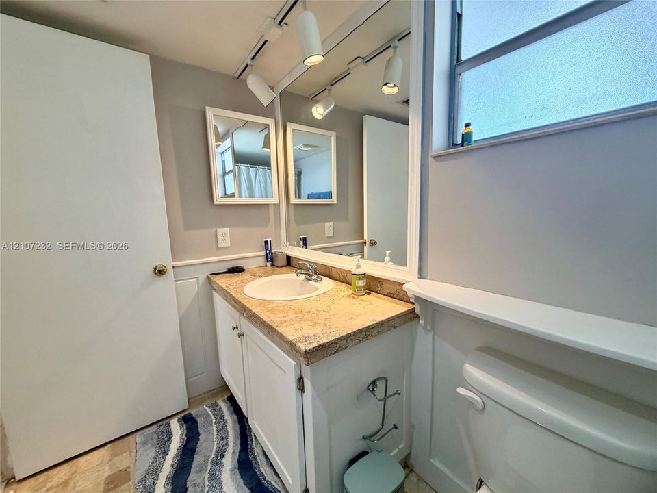 Vanity featuring a stone-look countertop with an integrated basin, white shaker-style cabinetry, wall-to-wall mirror, track lighting, and a window ledge