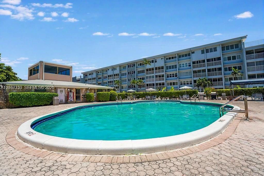 Oval-shaped swimming pool with a concrete surround and paved pool deck, featuring established perimeter landscaping and a stone-accented community building