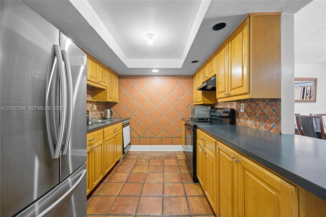 Kitchen featuring wood cabinetry, solid-surface countertops, terra-cotta tile flooring, patterned ceramic tile backsplash, and stainless-steel appliances
