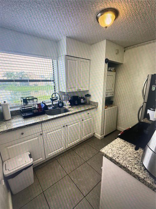 Kitchen featuring white cabinetry, stone-finish countertops, stainless steel sink with a pull-down faucet, large format tile flooring, and a stacked laundry unit