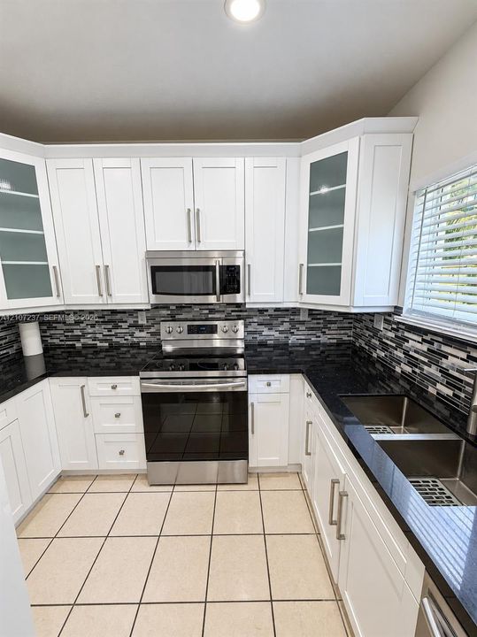 White shaker-style cabinetry with glass-front uppers, stainless steel appliances, dark stone countertops, mosaic tile backsplash, and dual-basin stainless steel sink