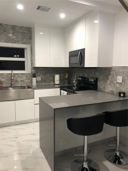 Modern kitchen featuring high-gloss white cabinetry, a solid surface waterfall-edge peninsula, stone-textured tile backsplash, stainless steel farmhouse sink, and recessed lighting