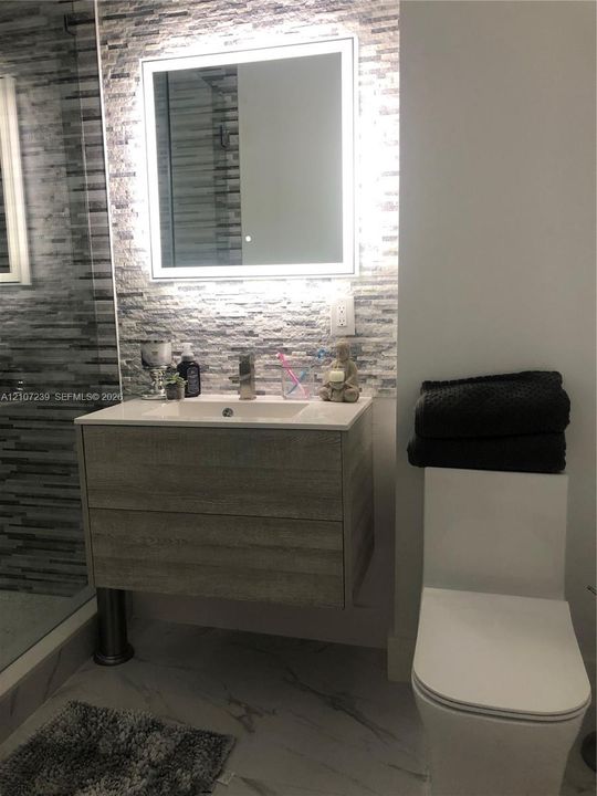Floating vanity with wood-finish cabinetry, rectangular integrated sink, LED-backlit mirror, stacked stone-texture wall accent, and porcelain tile flooring