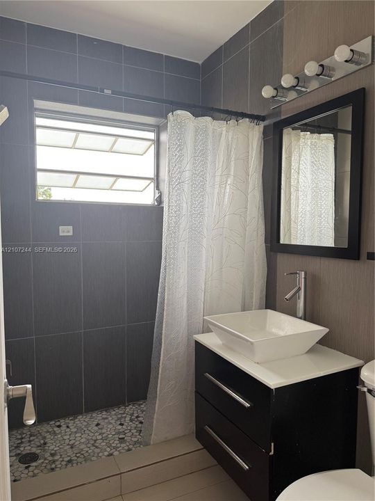 Bathroom featuring a floating vanity with a white vessel sink, dark-toned cabinetry, charcoal-hued wall tiling, pebble-texture shower flooring, and a framed mirror