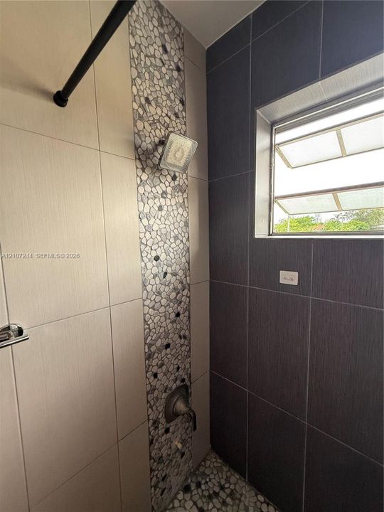 Shower enclosure featuring large-format cream and charcoal tile, a decorative vertical pebble-stone inlay, a pebble-stone floor, and a built-in window