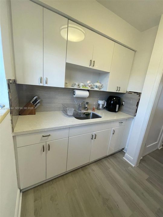 Glossy white cabinetry, stone-finish countertops, stainless steel undermount sink, horizontal tile backsplash, and light wood-finish flooring