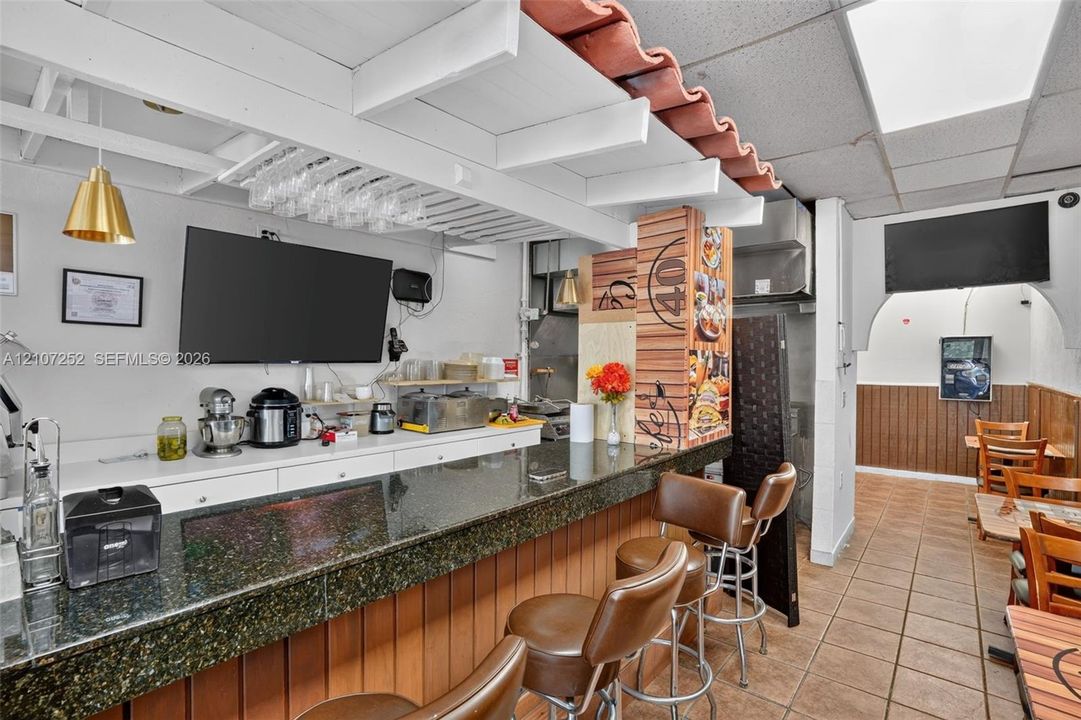 Built-in service bar featuring a solid surface countertop, wood-paneled base, decorative overhead beam structure, and integrated glass stemware rack