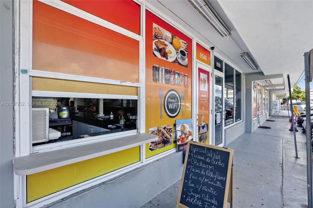 Commercial storefront exterior featuring a service window with a fixed counter, glass-paneled entry door, concrete walkway, and integrated linear lighting fixtures