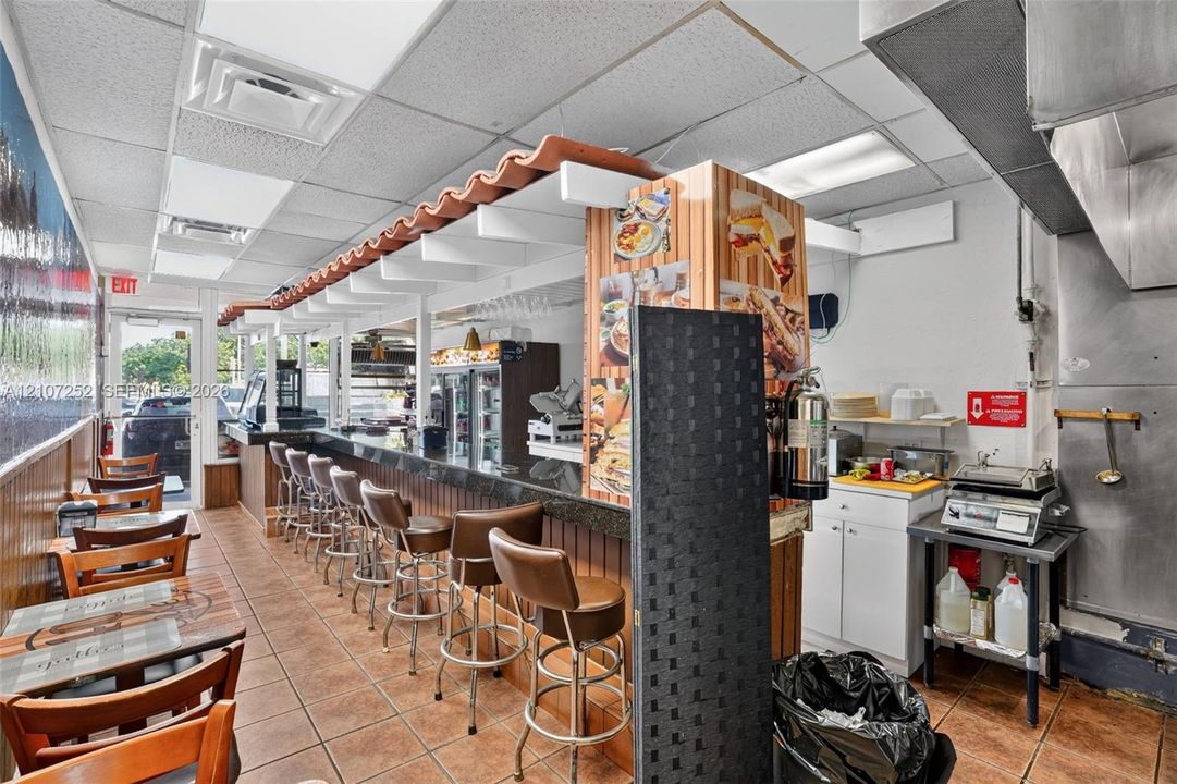 Commercial kitchen space featuring a wrap-around service counter, decorative tile-finish canopy, ceramic tile flooring, and integrated stainless steel exhaust ventilation