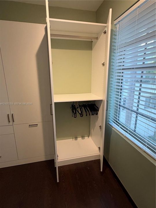 Built-in floor-to-ceiling cabinetry with white finish, integrated hanging rods, adjustable shelving, horizontal window blinds, and dark wood-finish flooring