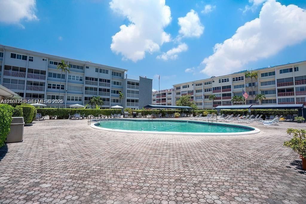 Oval swimming pool with a paved deck, surrounded by manicured hedges and covered seating areas
