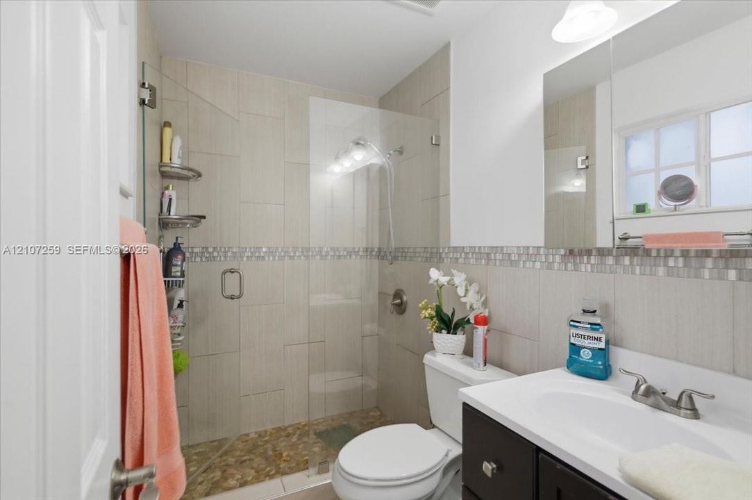Bathroom featuring a glass-enclosed shower with pebble-stone flooring, large-format neutral wall tile, decorative mosaic tile inlay, dark cabinetry with a white countertop, and a wall-mounted mirror