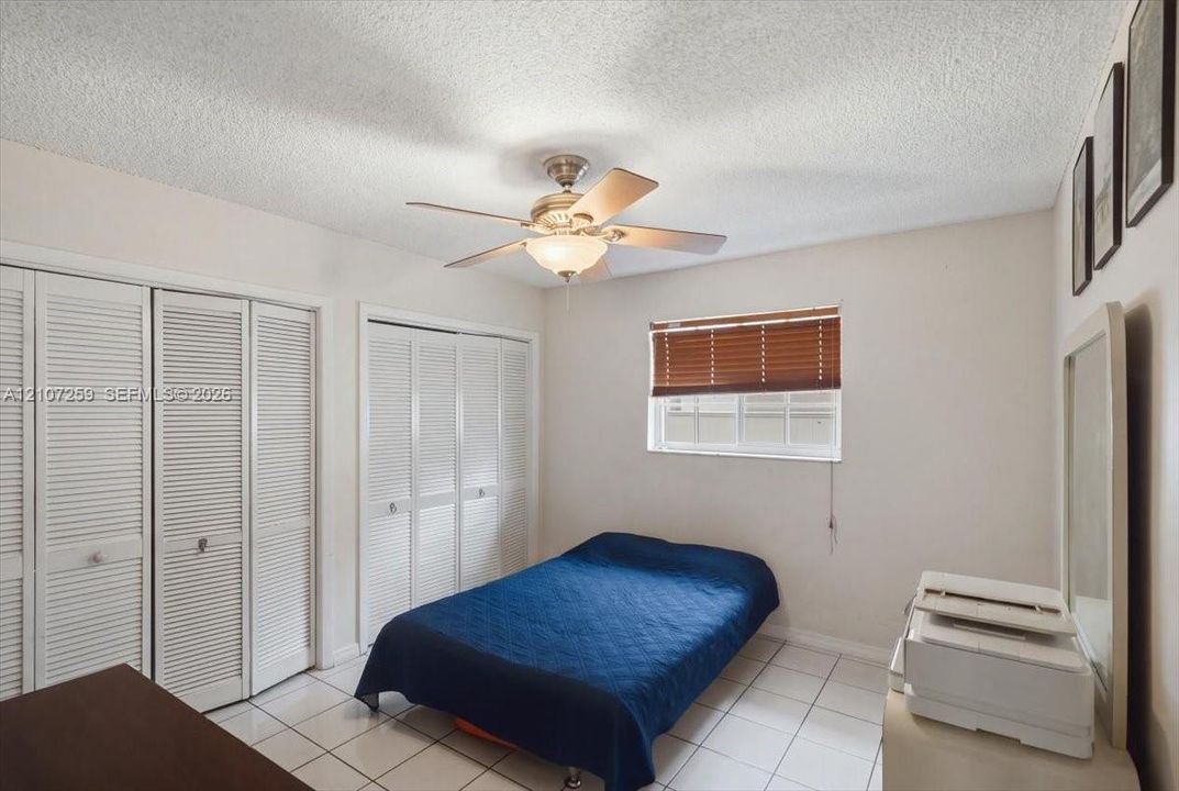 Neutral-toned room featuring light-colored floor tiling, dual louvered-door closets, a ceiling-mounted light fixture with integrated fan, and a window with horizontal blinds
