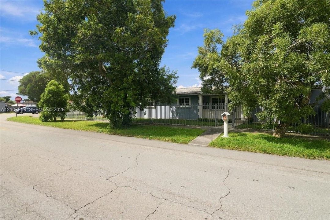 Single-story residence featuring a gray exterior, metal perimeter fencing, mature trees, and a concrete walkway