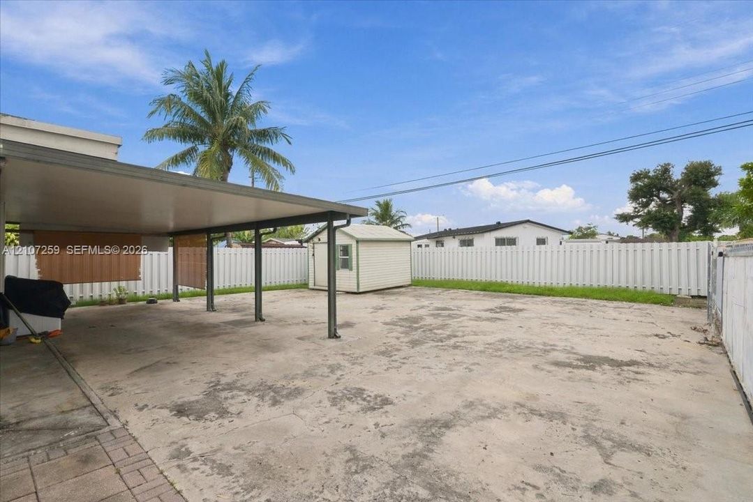 Expansive concrete courtyard featuring a covered carport, perimeter vinyl fencing, and a detached storage shed