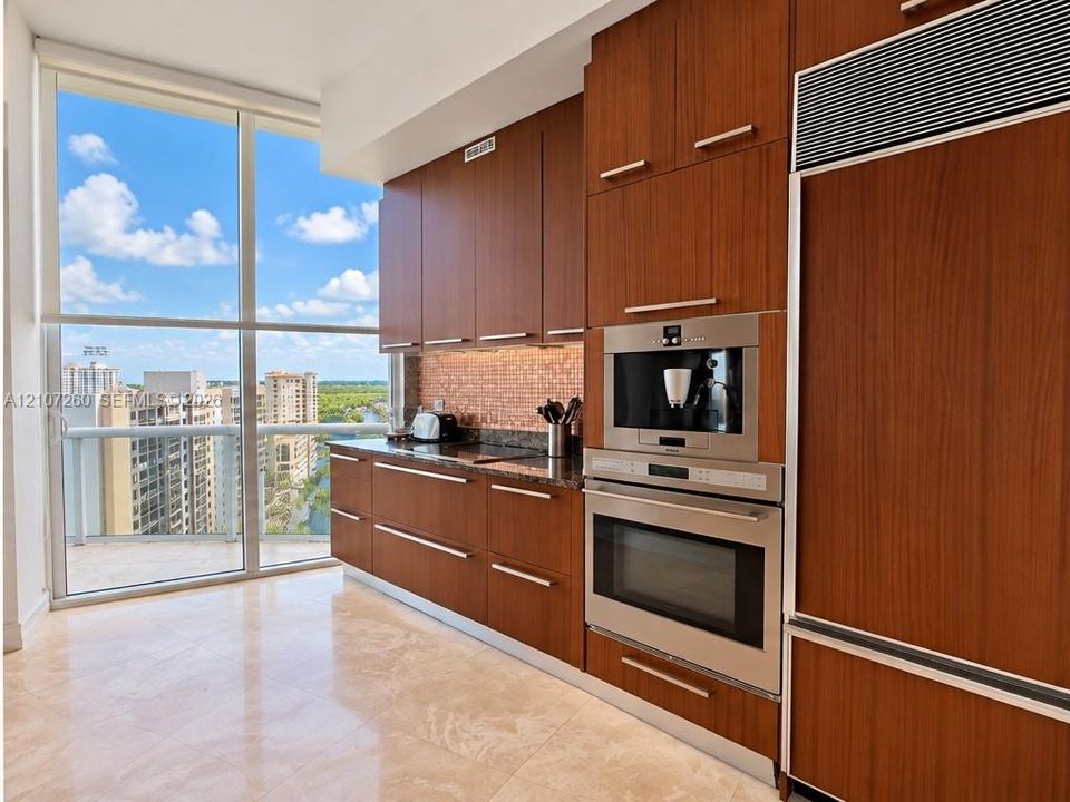 Kitchen featuring custom flat-panel wood cabinetry, integrated stainless steel refrigerator, built-in wall oven and coffee station, stone countertops with a mosaic tile backsplash, and floor-to-ceiling windows