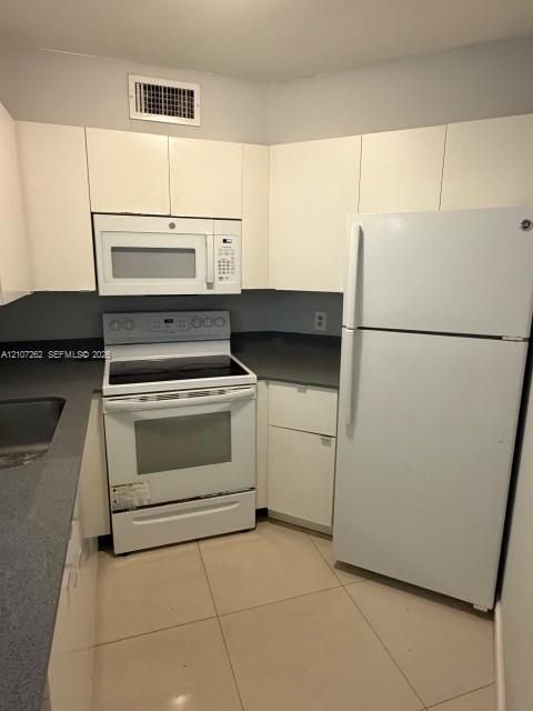 Kitchen featuring white flat-panel cabinetry, dark grey countertops, tile flooring, and white appliances including a built-in microwave, range, and refrigerator