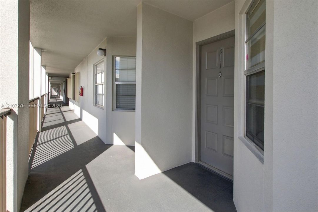 Exterior walkway featuring a six-panel door, neutral stucco walls, metal railings, and dual-pane windows