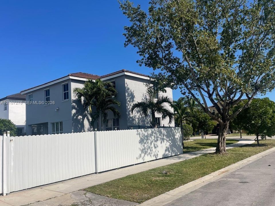 Two-story exterior with a hipped roof and grey stucco facade, enclosed by a white privacy fence and mature palm and shade trees